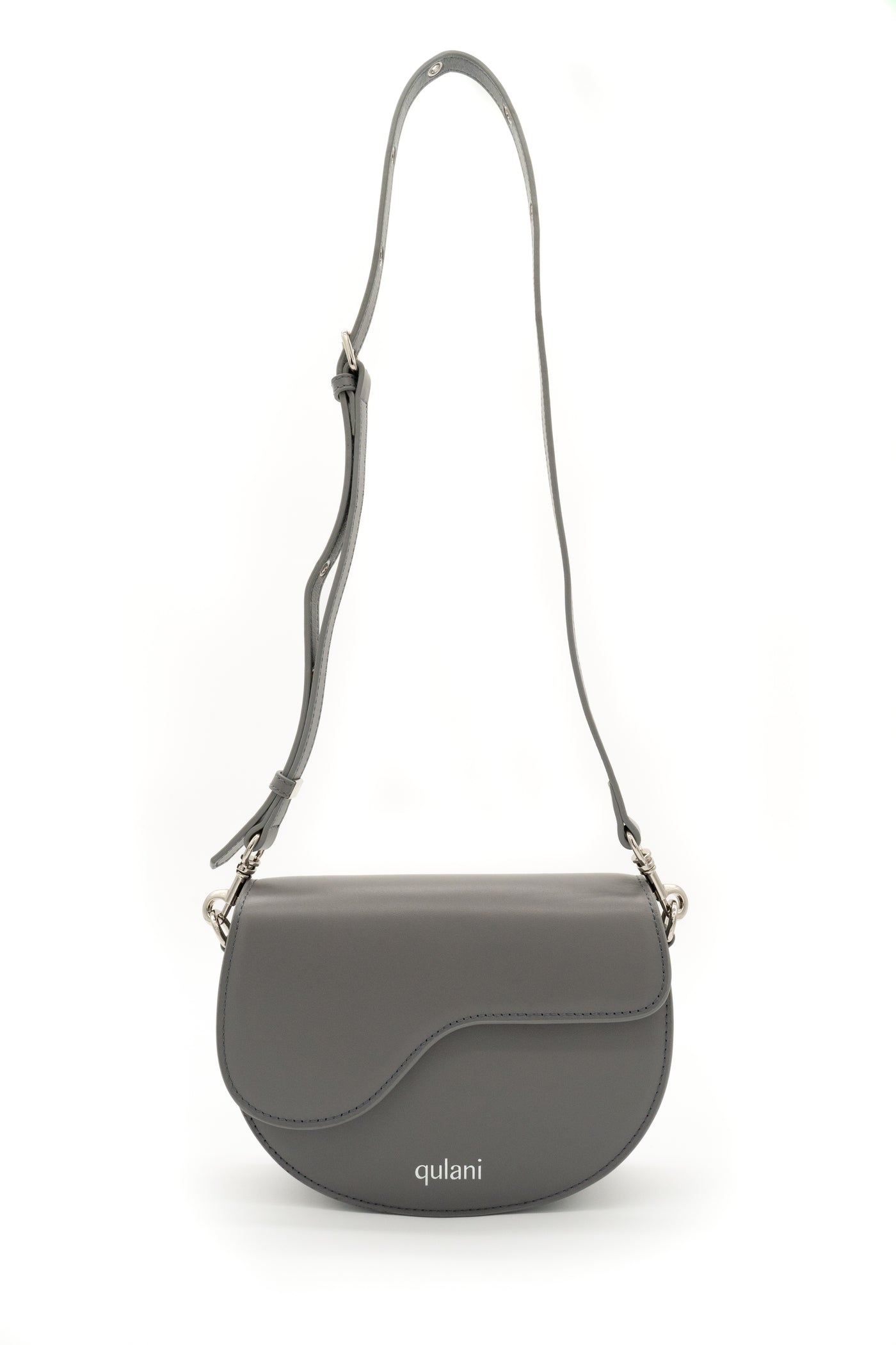 A gray leather Qulani shoulder saddle bag with a strap, featuring a front flap closure and a silver-tone logo on the front.