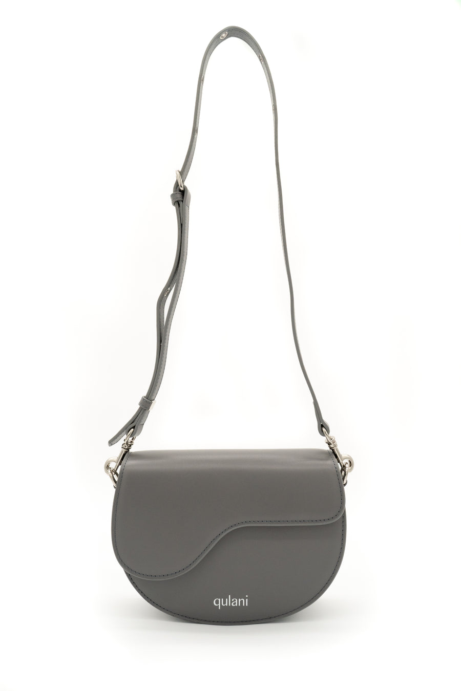 A gray leather Qulani shoulder saddle bag with a strap, featuring a front flap closure and a silver-tone logo on the front.